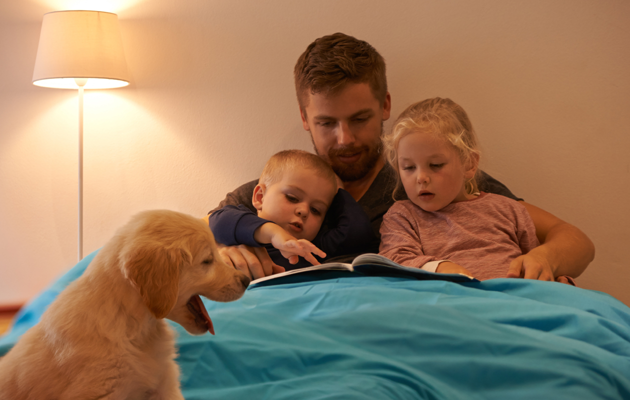 Isä lukee iltasatua sängyssä kahdelle lapselle, koira istuu etualalla.