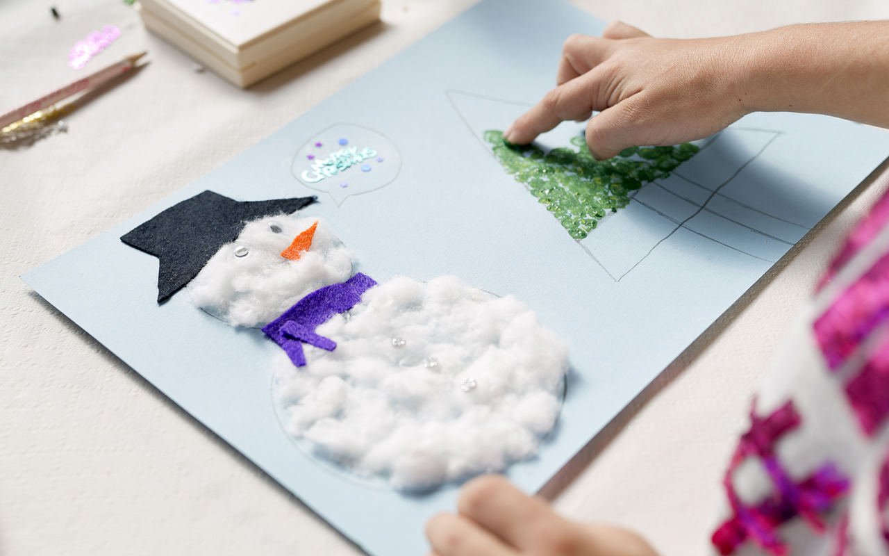 Joulukorttien askartelu lasten kanssa: lapsi maalaa lumiukon viereen kuusen.